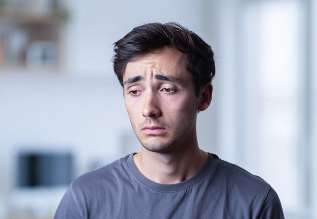 Молодой мужчина в тёмно-серой футболке стоит в светлой комнате с опущенным взглядом и грустным выражением лица